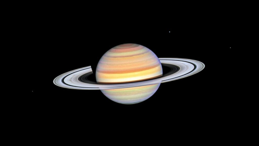 Planet Saturn with bright white rings, multi-colored main sphere, and moons Mimas, Dione, and Enceladus. Spoke features on the left and right sides of the rings appear like faint grey smudges against the rings’ bright backdrop, about midway from the planet to the rings’ outer edge. Above the rings plane, the planet’s bands are shades of red, orange and yellow, with bright white nearer the equator.