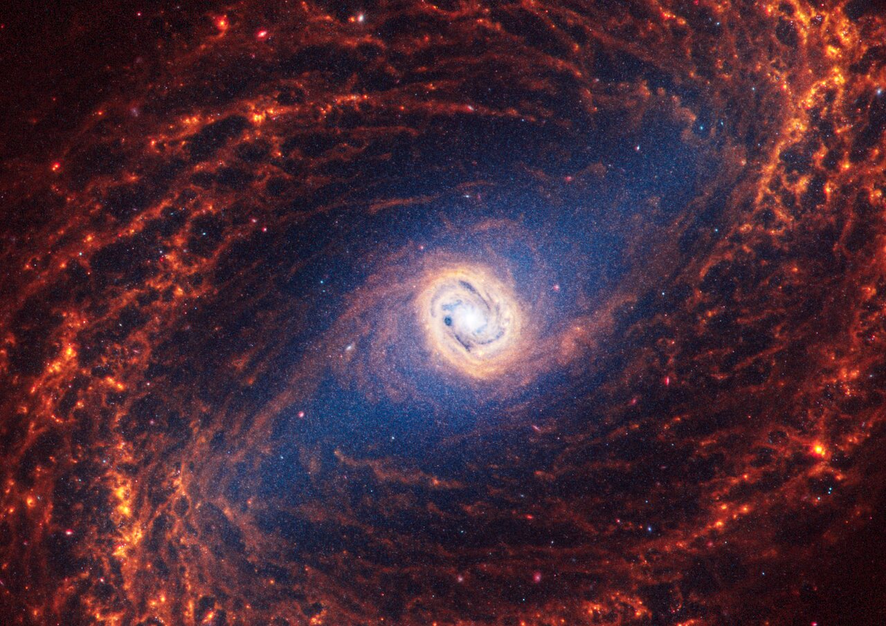Webb’s image of NGC 1433 shows a face-on barred spiral galaxy anchored by its central region, which is circular and shows a bright white point at the centre with a light yellow circle around it. The galaxy has a large bar connected to filamentary spiral orange arms that appear to rotate counterclockwise.