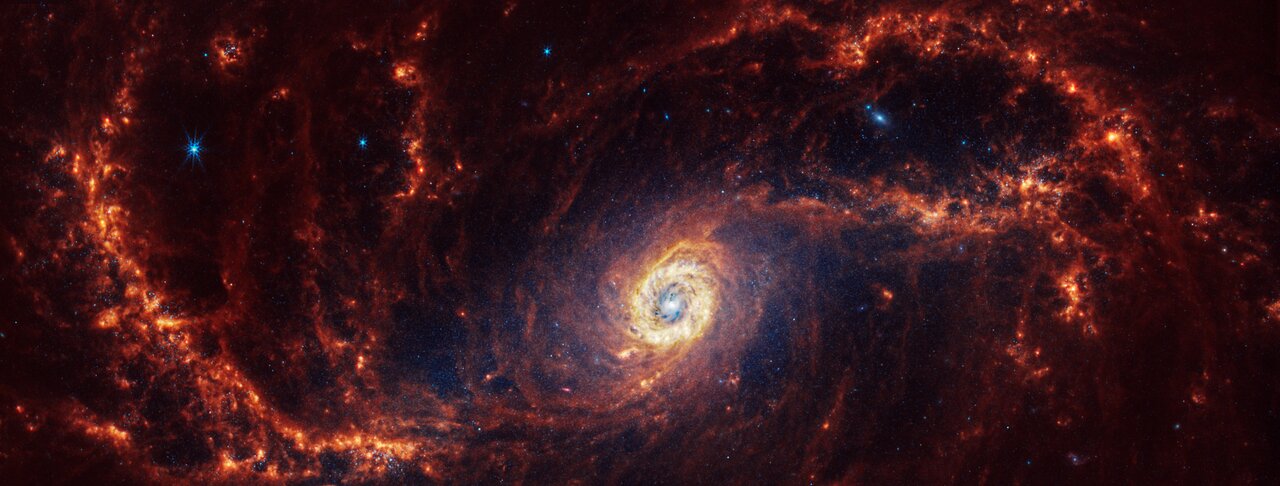Webb’s image of NGC 1672 shows a portion of a densely populated face-on spiral galaxy anchored by a central core and bar structure made of filamentary dust lanes and a light blue haze of stars, just below centre. Two spiny spiral orange arms extend from the bar to the edges and rotate clockwise.