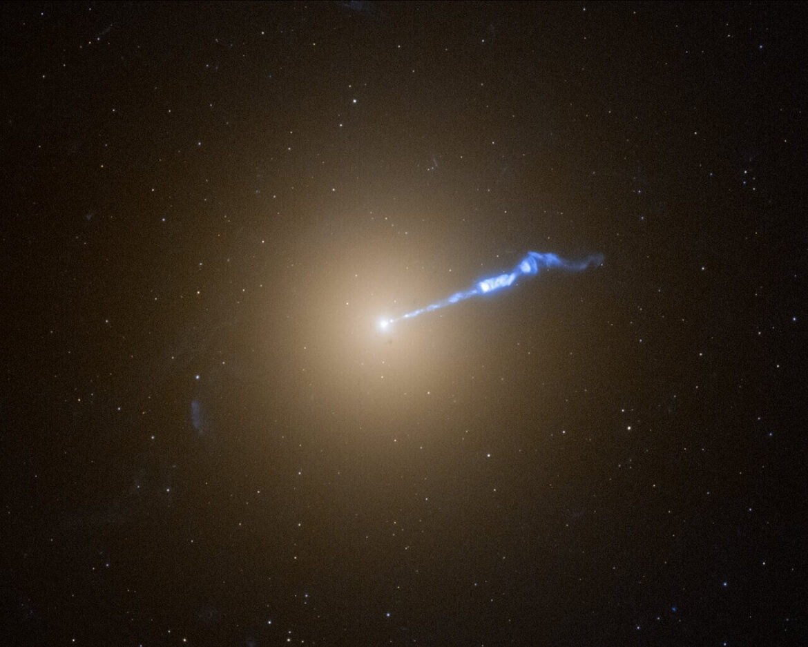 A Hubble photo of galaxy M87, which resembles a translucent, fuzzy white cotton ball. The brightness decreases gradually out in all directions from a bright white point of light at the centre. A wavy blue-white jet of material extends from the point-like core outward to the upper right, about halfway across the galaxy. Stars speckle the background.