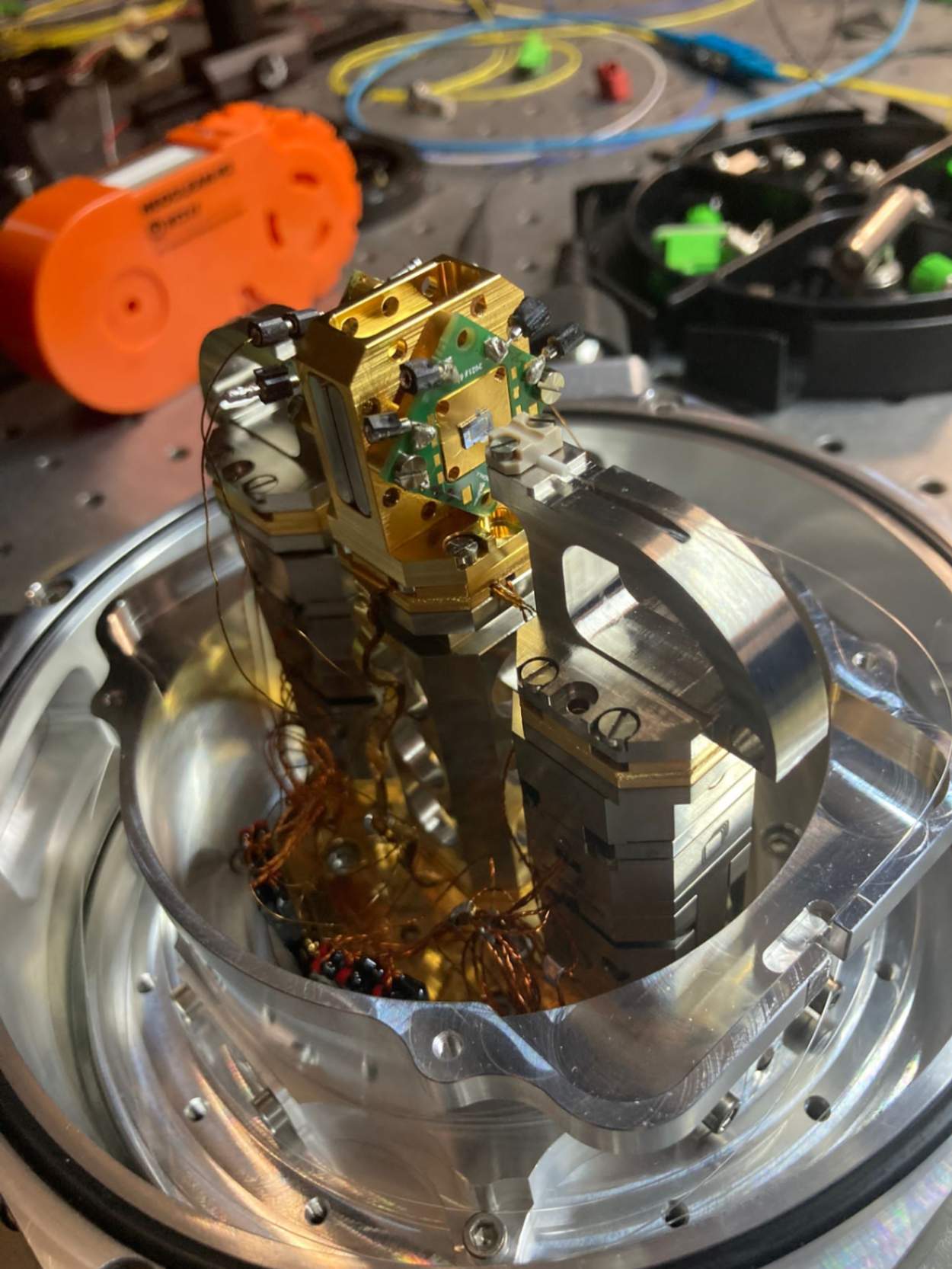 Interno della camera del criostato che ospita il campione contenente la sorgente quantum dot Quandela. Nella fotografia si può osservare il campione (rettangolo grigio circondato esternamente prima dal quadrato d’oro e poi dal rombo verde) e l’alloggio della fibra che colleziona i singoli fotoni emessi, posizionata su traslatori piezoelettrici (parte in grigio)