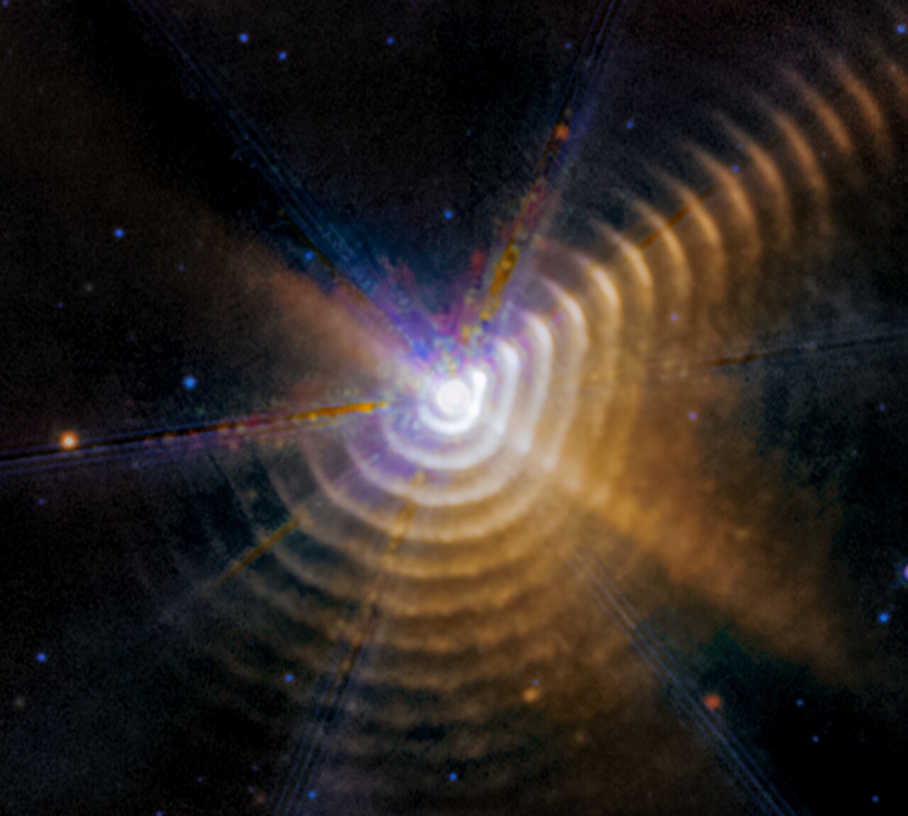 The image shows a bright white point of light surrounded by 17 regularly spaced, hazy dust shells at the bottom, right, and upper right, which look like tree rings. There is noticeably less color in the upper left. The central point, where the two stars are located, has a roughly hexagonal shape.