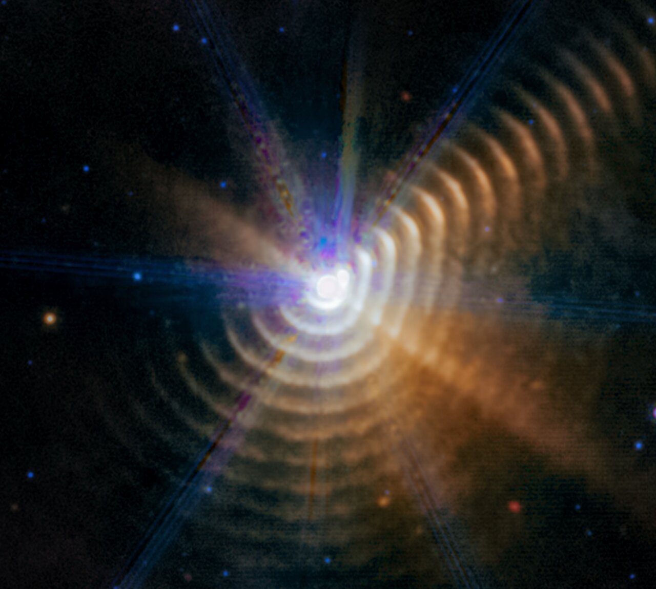 The image shows a bright white point of light surrounded by 17 regularly spaced, hazy dust shells at the bottom, right, and upper right, which look like tree rings. There is noticeably less color in the upper left. The central point, where the two stars are located, has a roughly hexagonal shape.