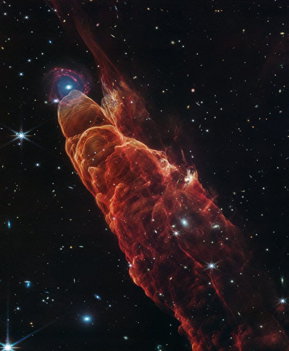 Angled from the upper left corner to the lower right corner of the image is a conical shaped orange-red cloud known at Herbig-Haro 49/50. This feature takes up about three-fourths of the length of this angle. The upper left end of this feature has a translucent, rounded end. At this same location there is a background spiral shaped galaxy with a concentrated blue center that fades outwards to blend in with red spiral arms. The conical feature widens slightly from the rounded end at the upper right down to the lower right. The black background of space is clearer, speckled with some white stars and smaller, more numerous, fainter white galaxies.